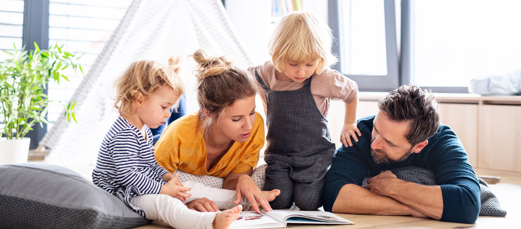 Front view of young family with two small children indoors in bedroom reading a book.